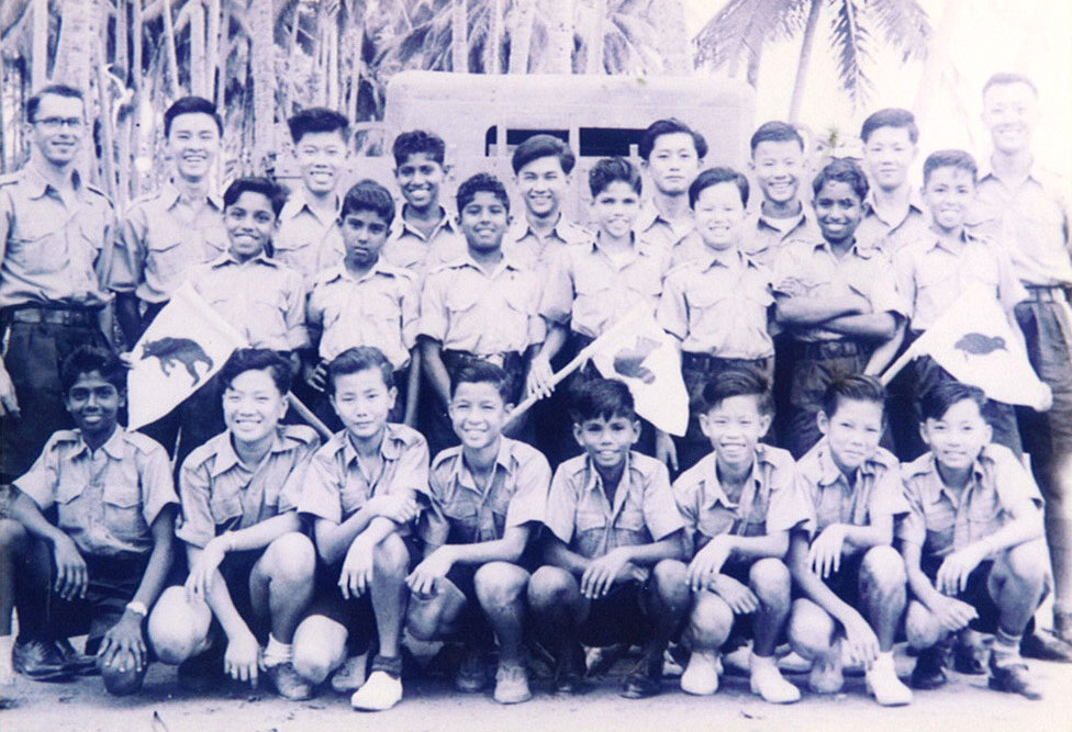 A vintage black-and-white group photo of boys in uniform posing outdoors near palm trees and a vehicle.